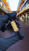 Santa Muerte gold charm and black braided nylon bracelet held in a gloved hand